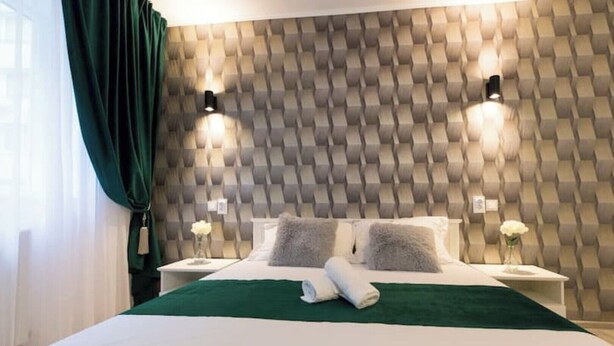 Ein ordentlich bezogenes Bett mit einem grünen Bettüberwurf, zwei Kissen und gerollten Handtüchern auf einer weißen Matratze. Zwei Nachttische mit Lampen und eine Wand mit geometrischem Muster sind zu sehen.