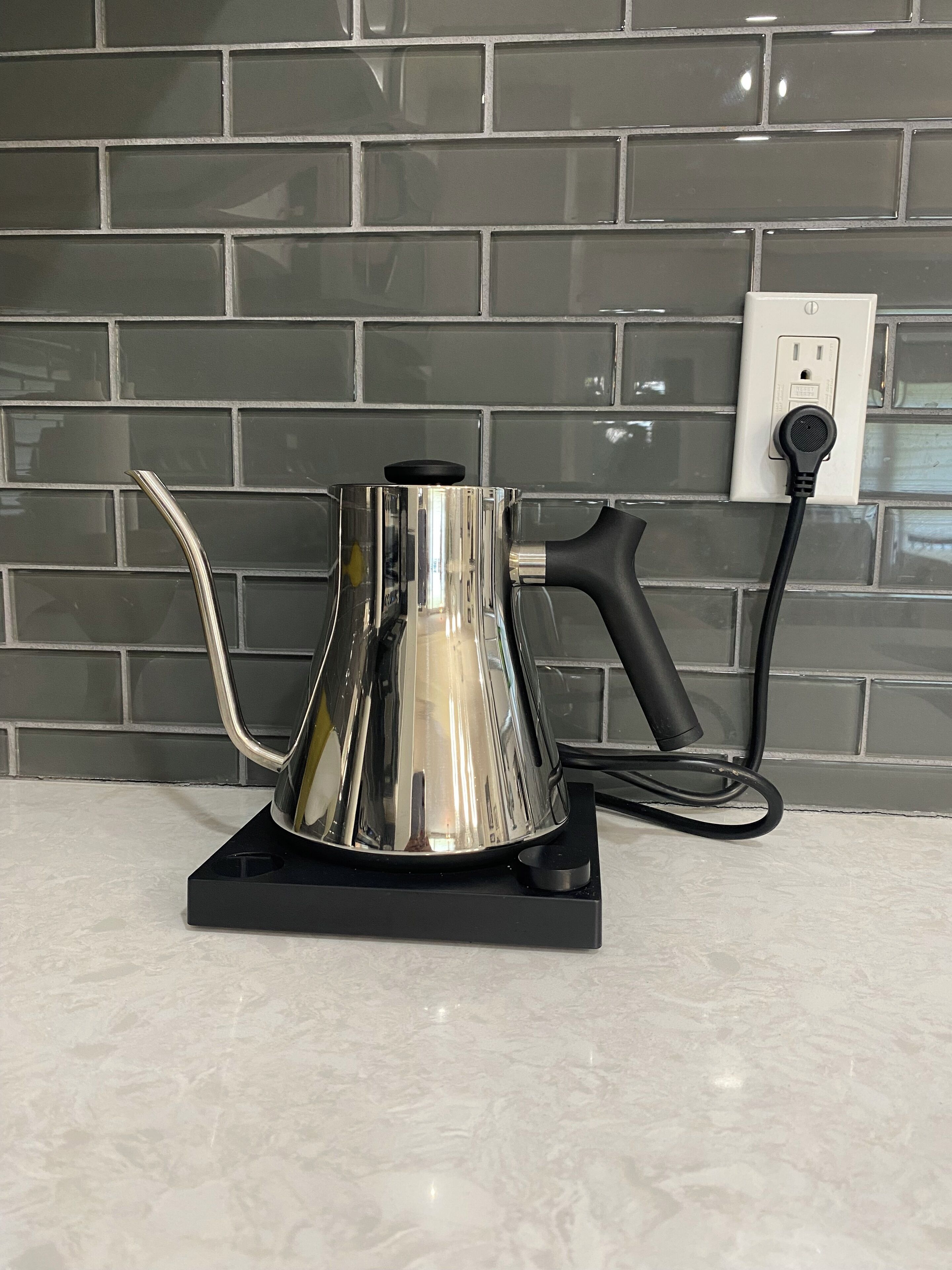Eine Küche mit einer Kaffeemaschine auf einem schwarzen Standfuß, einer Steckdose und einer gefliesten Spritzschutzwand.