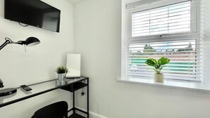 Una habitación pequeña y moderna con escritorio, silla y televisor montado en la pared. Hay una ventana con persianas y una planta en maceta en el alféizar.