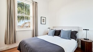 A bedroom with a bed, a nightstand, a lamp, a window with curtains, and a framed picture on the wall. - Coogee Treetops ISYD (Coogee)