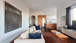A modern living room with a sofa, coffee table, and artwork. A kitchen is visible in the background with a refrigerator and cabinets. - Coogee Treetops ISYD (Coogee)