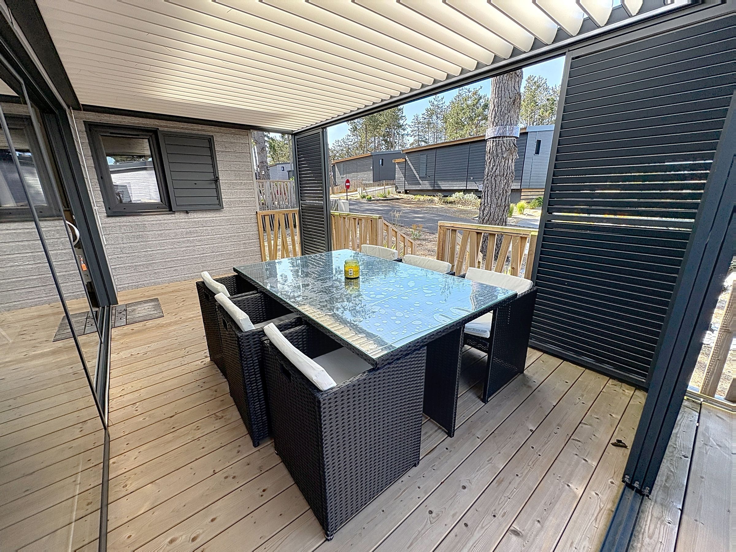 Een terras buiten met een tafel met glazen blad en rieten stoelen, omringd door houten terrasplanken en een metalen hekwerk.