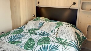 Un lit avec une couvre-lit orné d’un motif de feuilles tropicales, une tête de lit sombre et une armoire en bois avec une découpe circulaire.