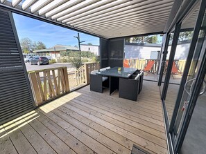 Una terraza techada al aire libre con una mesa y sillas, baranda de madera y vista a la calle.