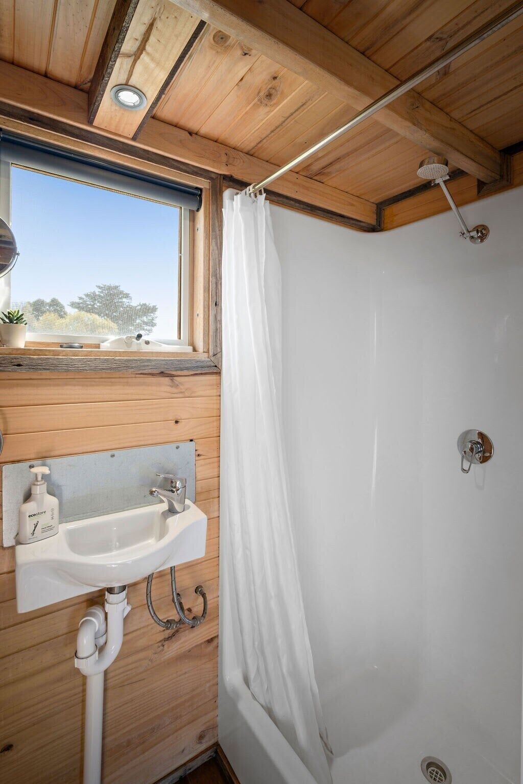 Une salle de bain rustique, aménagée dans une cabane en bois, avec une fenêtre, un lavabo et une douche.