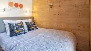 A bedroom with a wooden ceiling, a bed with white bedding and patterned pillows, a fluffy white rug, and a wall-mounted light fixture.