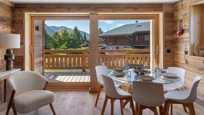 Zona pranzo con tavolo in vetro, sedie bianche e vista sulle montagne attraverso ampie porte scorrevoli.