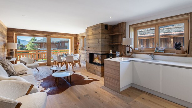Un interno moderno in stile baita con zona pranzo, camino, cucina e ampie finestre che offrono vista sulle montagne.