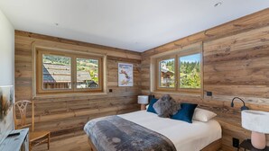 Una camera da letto con pareti in legno, un letto con coperta a motivi decorativi, due finestre che si affacciano su un villaggio e un piccolo tavolo con una lampada.