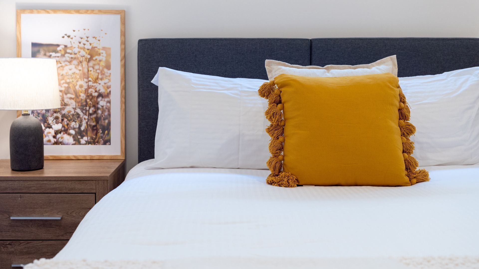 A neatly made bed with white linens and a mustard-colored pillow with tassels.