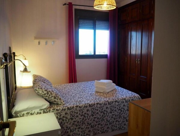 Ein Schlafzimmer mit Bett, Nachttischlampe, Fenster mit Vorhängen und einem Kleiderschrank.
