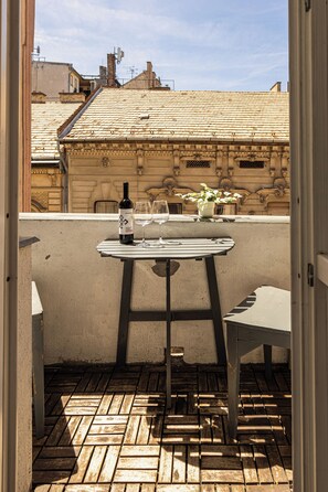 A balcony with a table, a bottle of wine, and glasses, overlooking a building with a tiled roof.