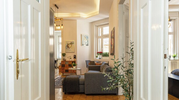 Um corredor com piso de madeira em padrão herringbone, portas brancas e uma sala de estar com sofá e planta em vaso.