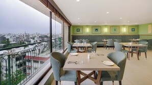 Un comedor con mesas y sillas, vista de la ciudad a través de amplios ventanales y una pared verde con cuadros enmarcados.