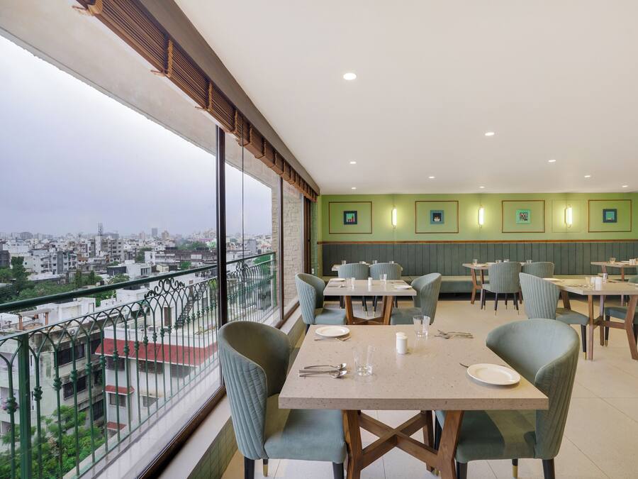 Un comedor con mesas y sillas, vista de la ciudad a través de amplios ventanales y una pared verde con cuadros enmarcados.