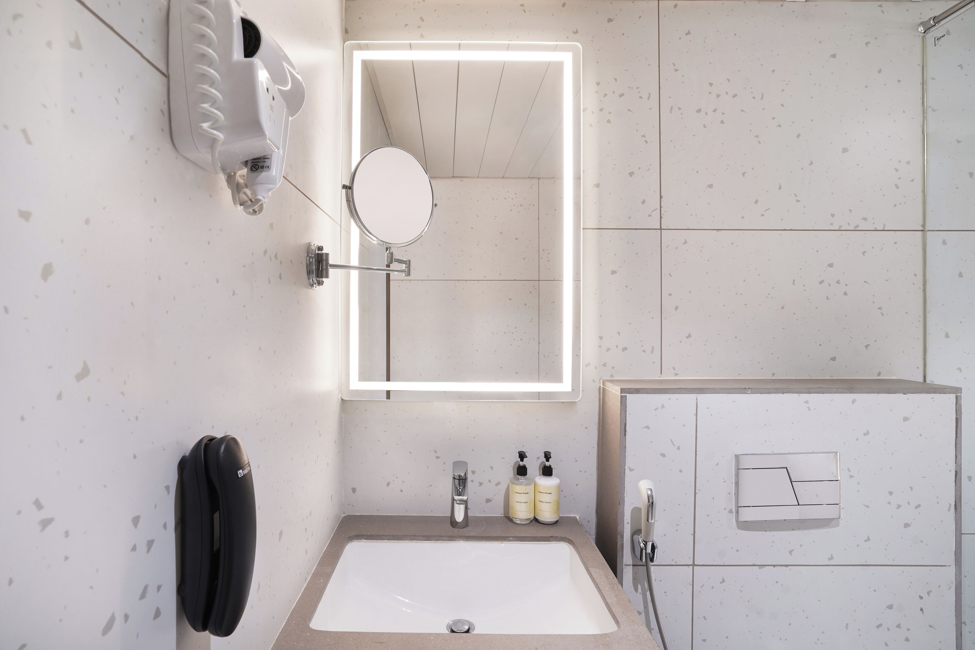 A bathroom with a white sink, a mirror with built-in lighting, a towel dispenser, and a shower area.