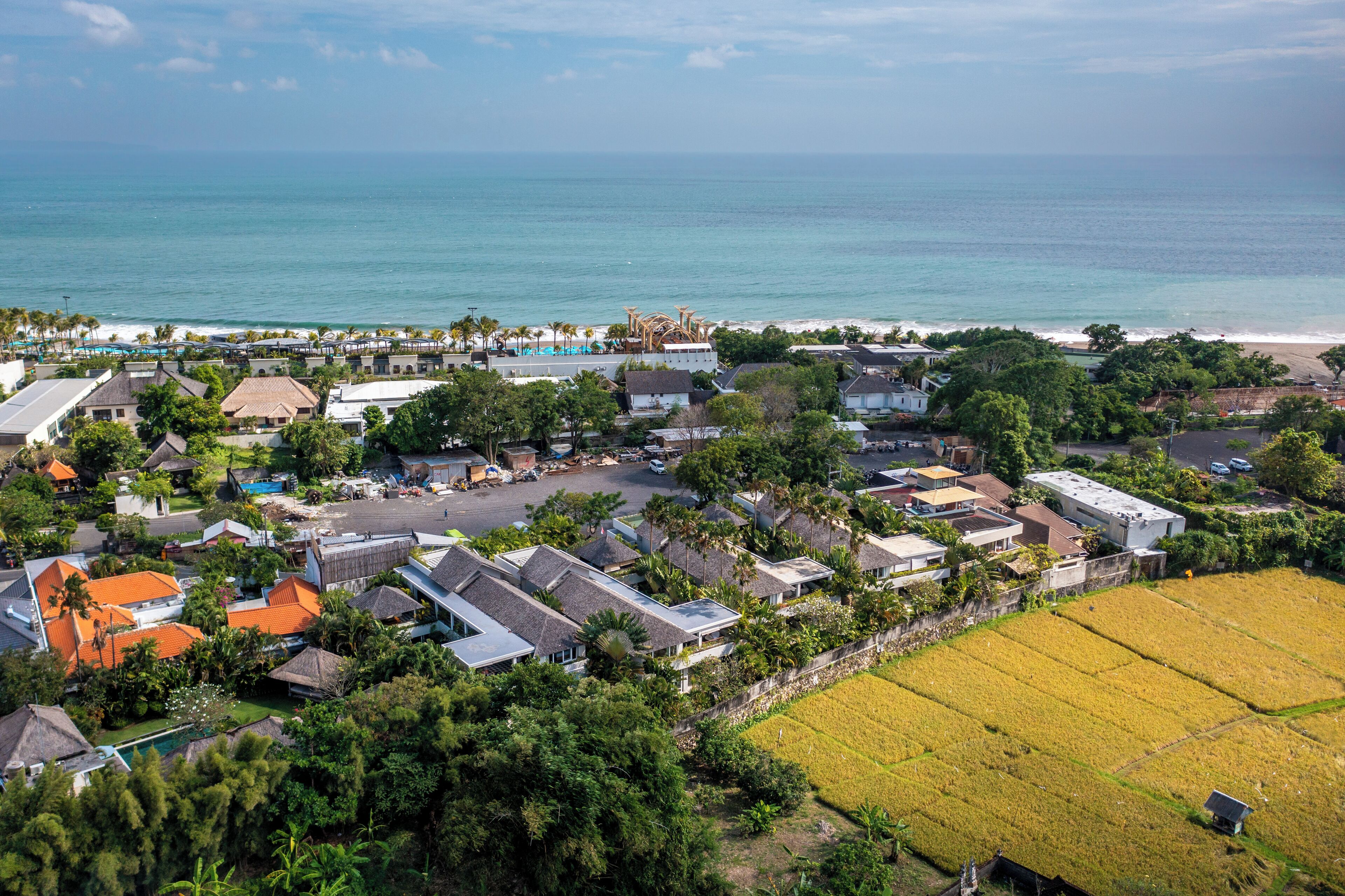 An aerial view of a coastal residential area with houses, a beach, and a large rice field.