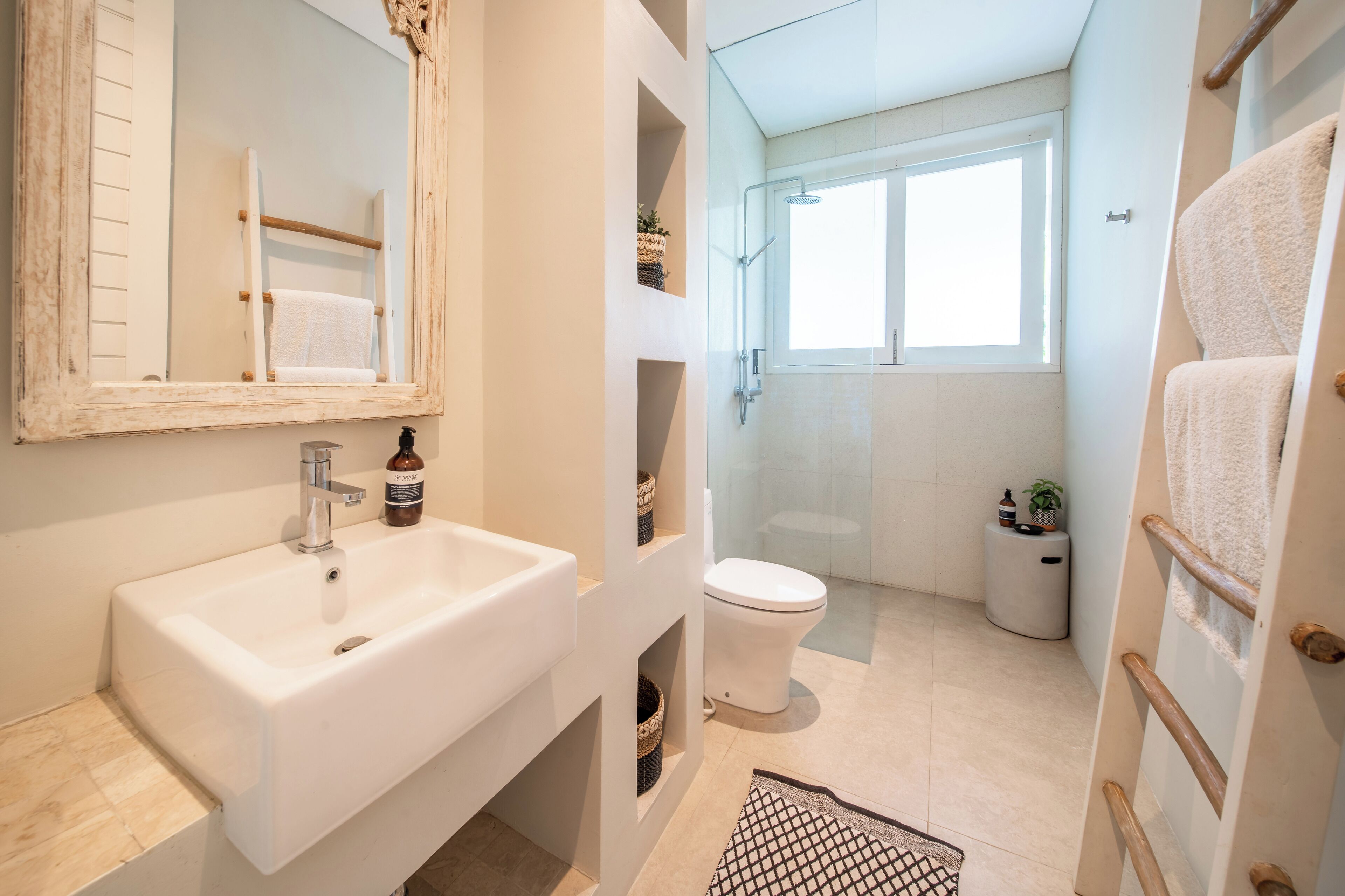 A bathroom with a white sink, mirror, and toilet. There is a window providing natural light and a small plant on the floor.