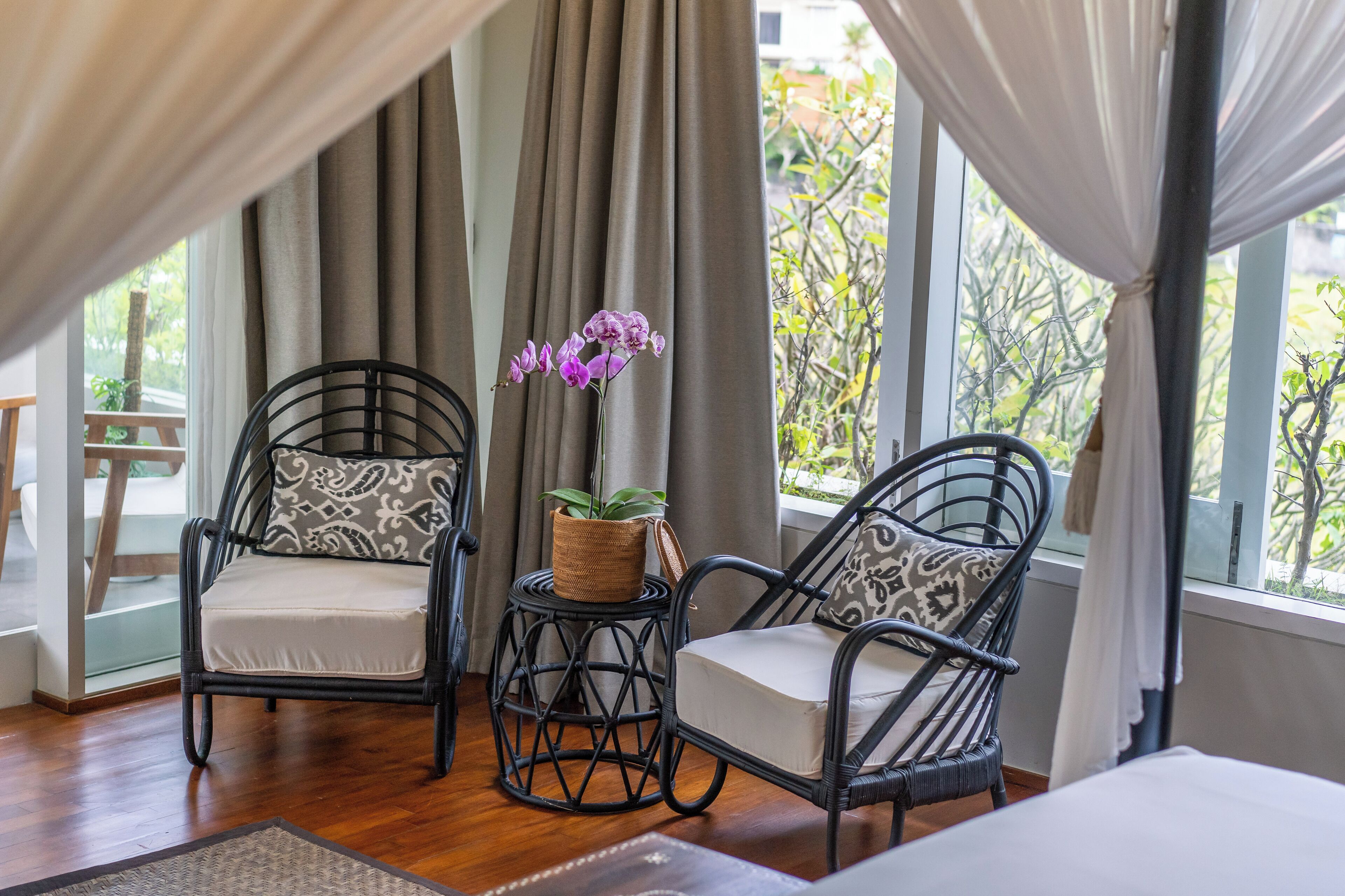 A room with two wicker chairs, a small round table with a plant, and a view of greenery outside.