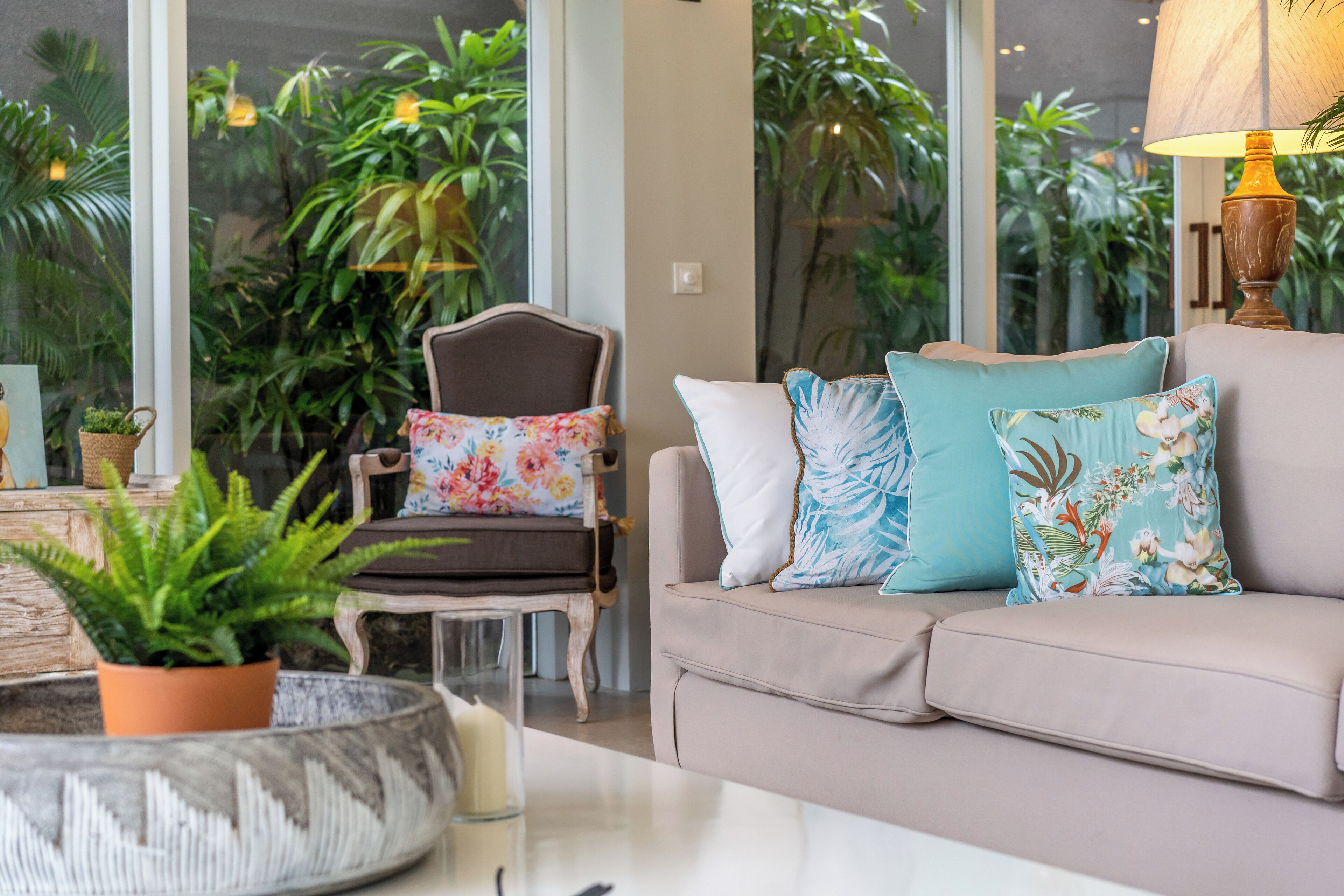 A cozy living room with a beige sofa, floral cushions, and a chair with a floral pillow. There is a potted plant on a table and a lamp on a side table.