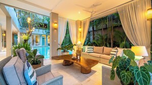 A modern interior with a wooden coffee table, wicker furniture, and a view of a pool and greenery. - Villa Sunsara 4  (Canggu)