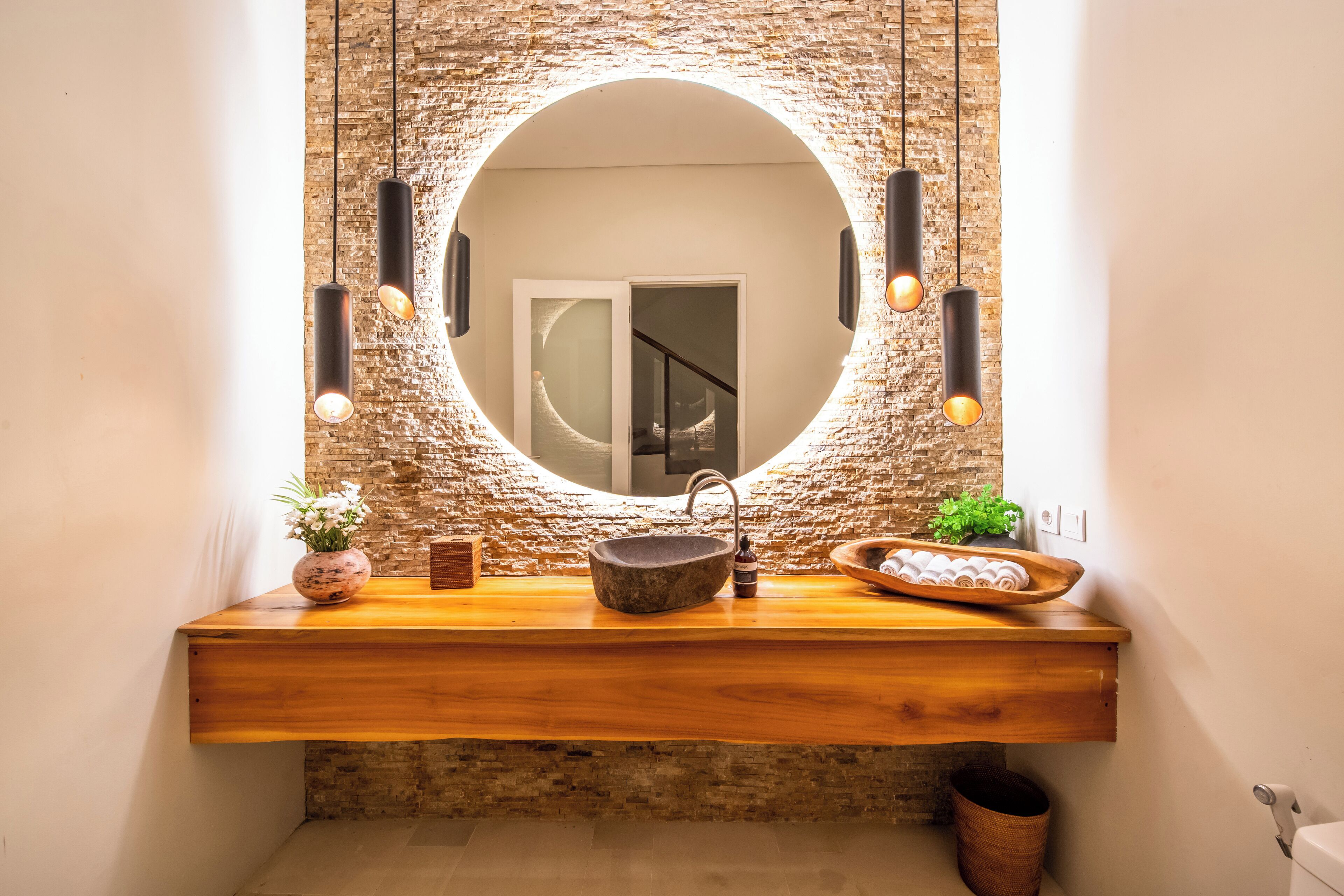 A bathroom with a round mirror, wooden countertop, stone sink, hanging pendant lights, and a view of a staircase.