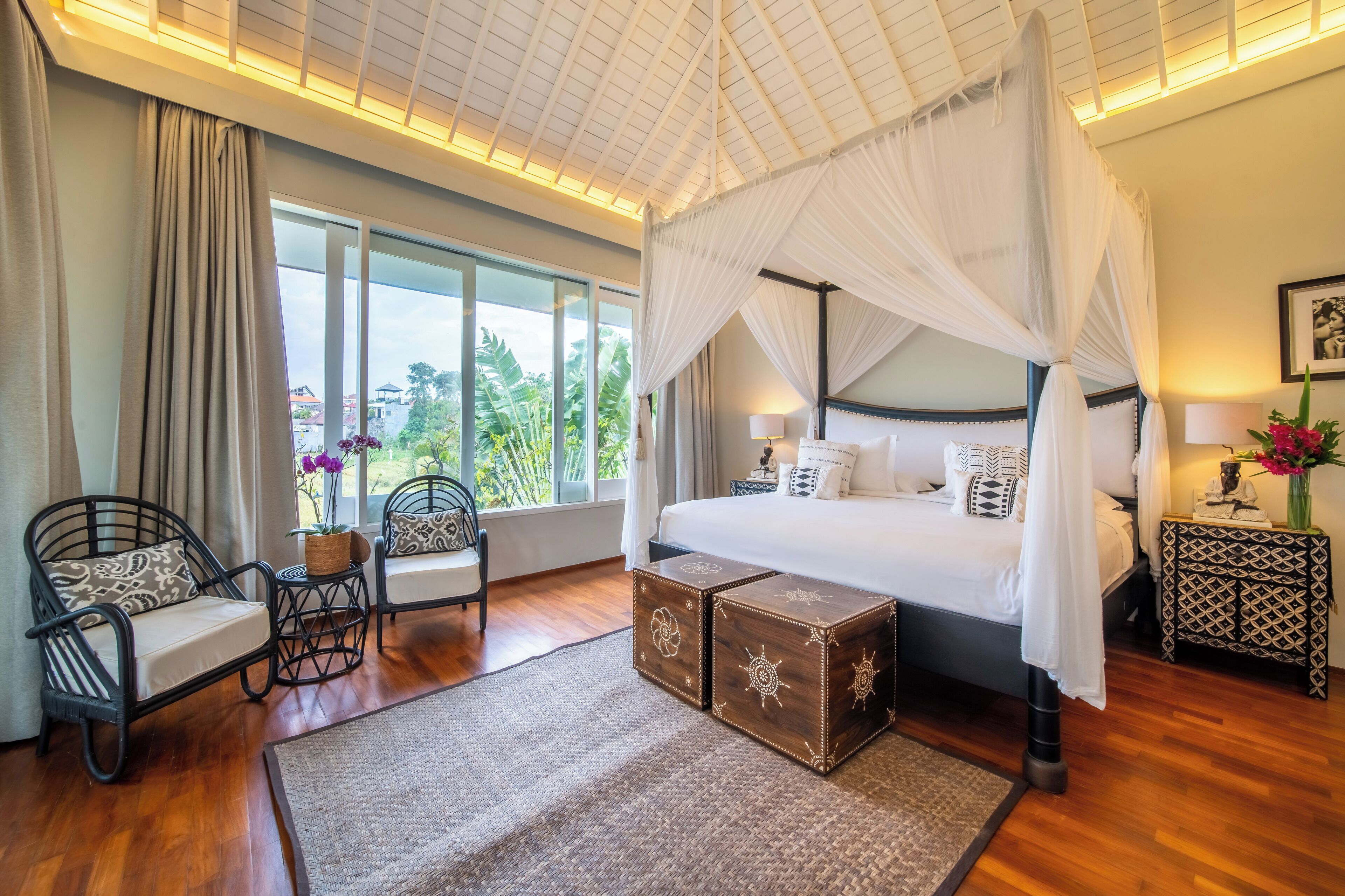 A bedroom with a four-poster bed, a sitting area with chairs, a small table, and a nightstand with a lamp.