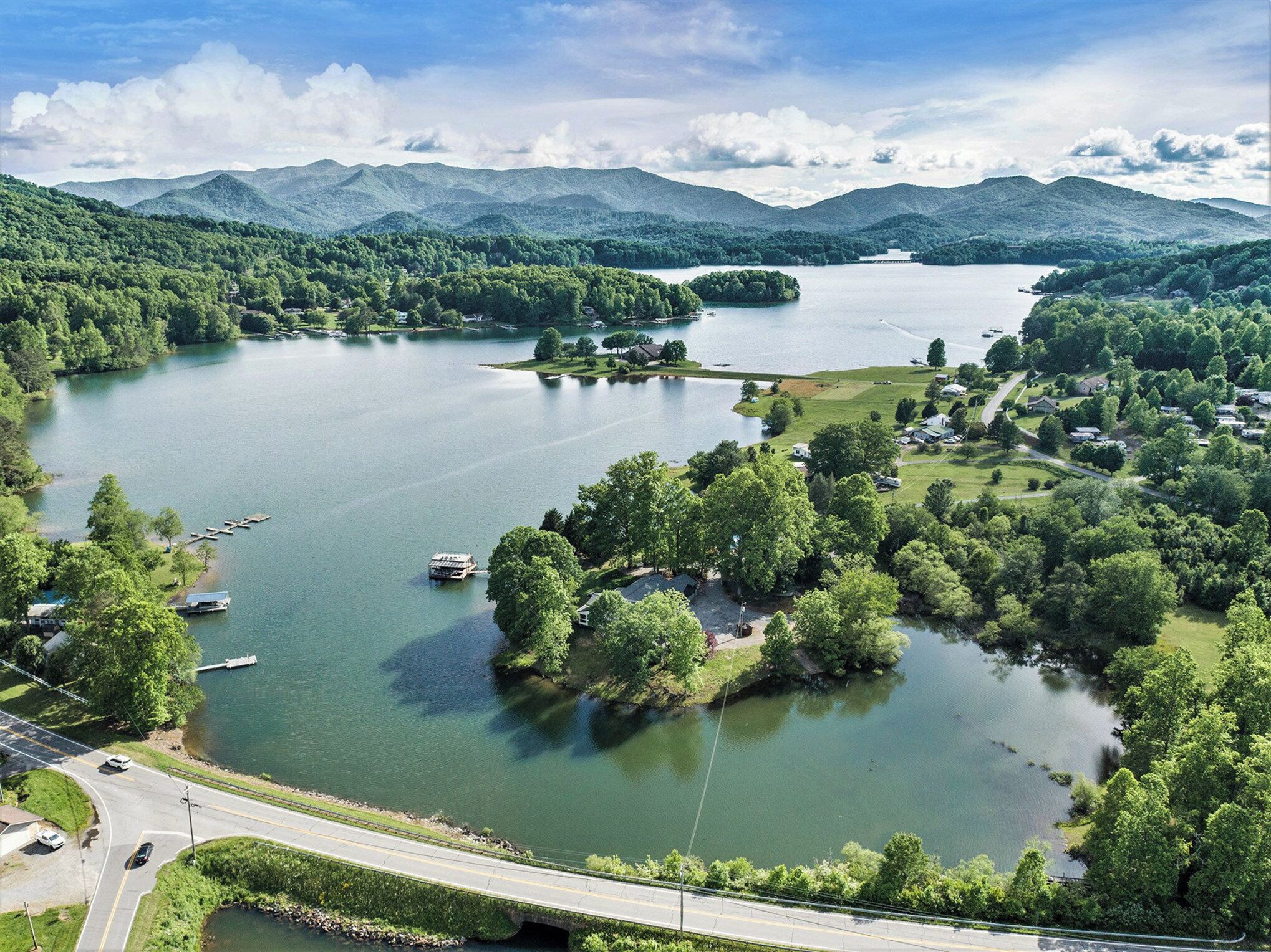 Un lago rodeado de exuberante vegetación y montañas. Hay barcos amarrados cerca de la orilla, y una carretera corre a lo largo de la orilla del lago.