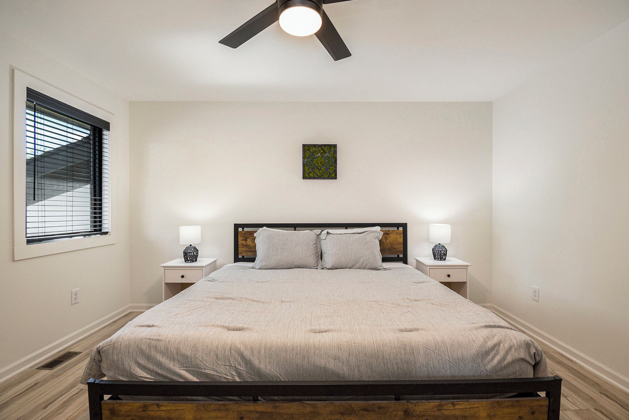 Une chambre à coucher comprenant un lit, deux tables de chevet avec des lampes, un ventilateur de plafond, une fenêtre avec des stores et une œuvre d’art accrochée au mur.