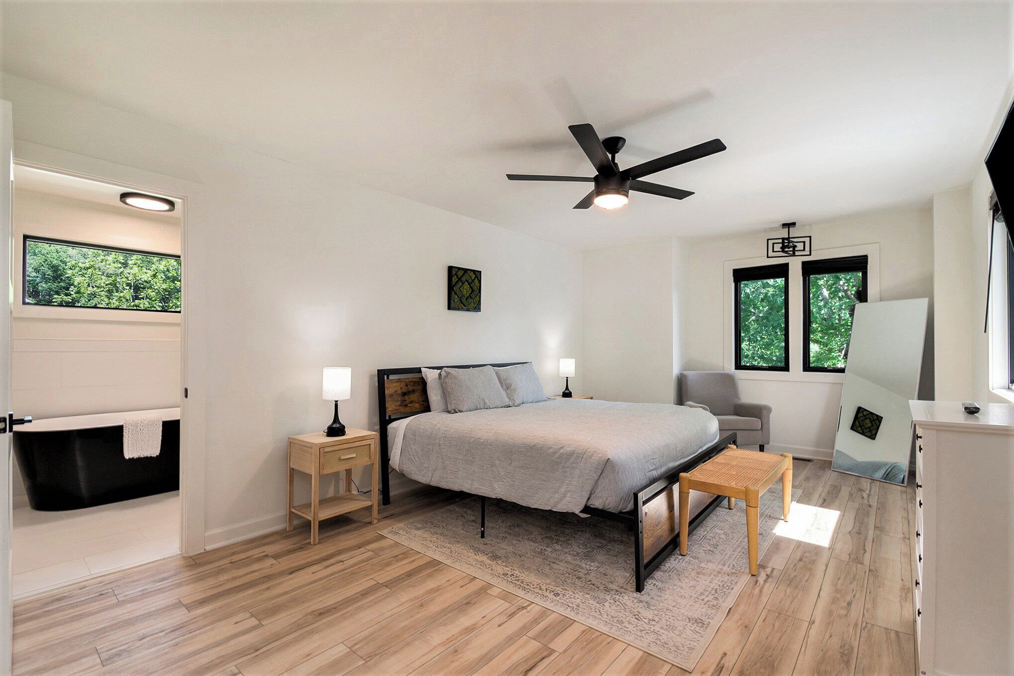 Un dormitorio moderno con cama, mesitas de noche, ventilador de techo, ventana y bañera integrada.