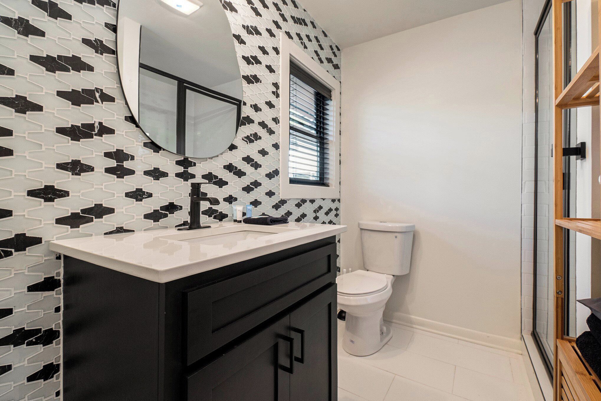 Une salle de bain avec un mur orné d’un motif noir et blanc, un lavabo blanc avec un robinet noir, des toilettes blanches et une fenêtre avec des stores.