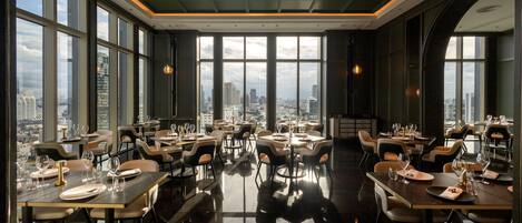 A modern restaurant with a panoramic city view, featuring high ceilings, large windows, and contemporary furniture.