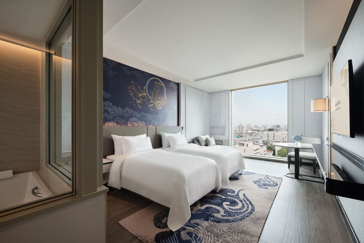 A modern hotel room with two beds, a large window with a city view, a dining area, and a flat-screen TV.