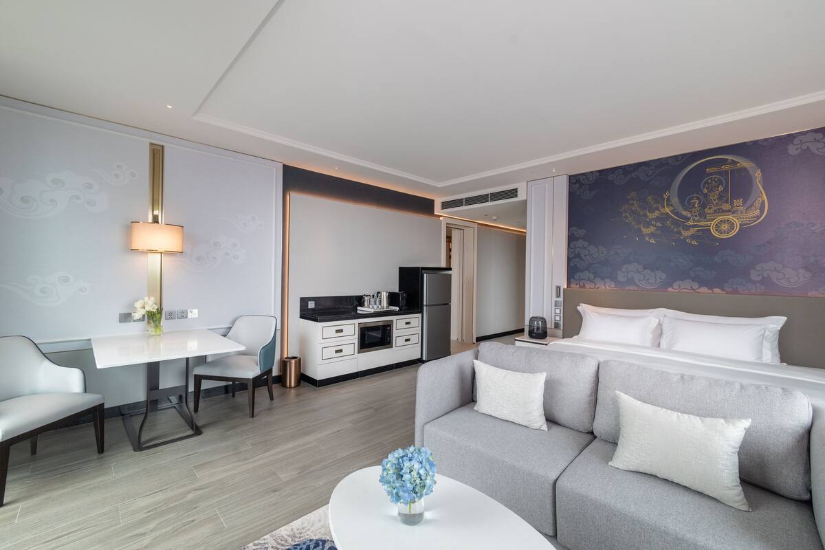 A modern hotel room with a grey sofa, a white coffee table, a dining area with a white table and chairs, a kitchen area, and a decorative wall with a golden car design.
