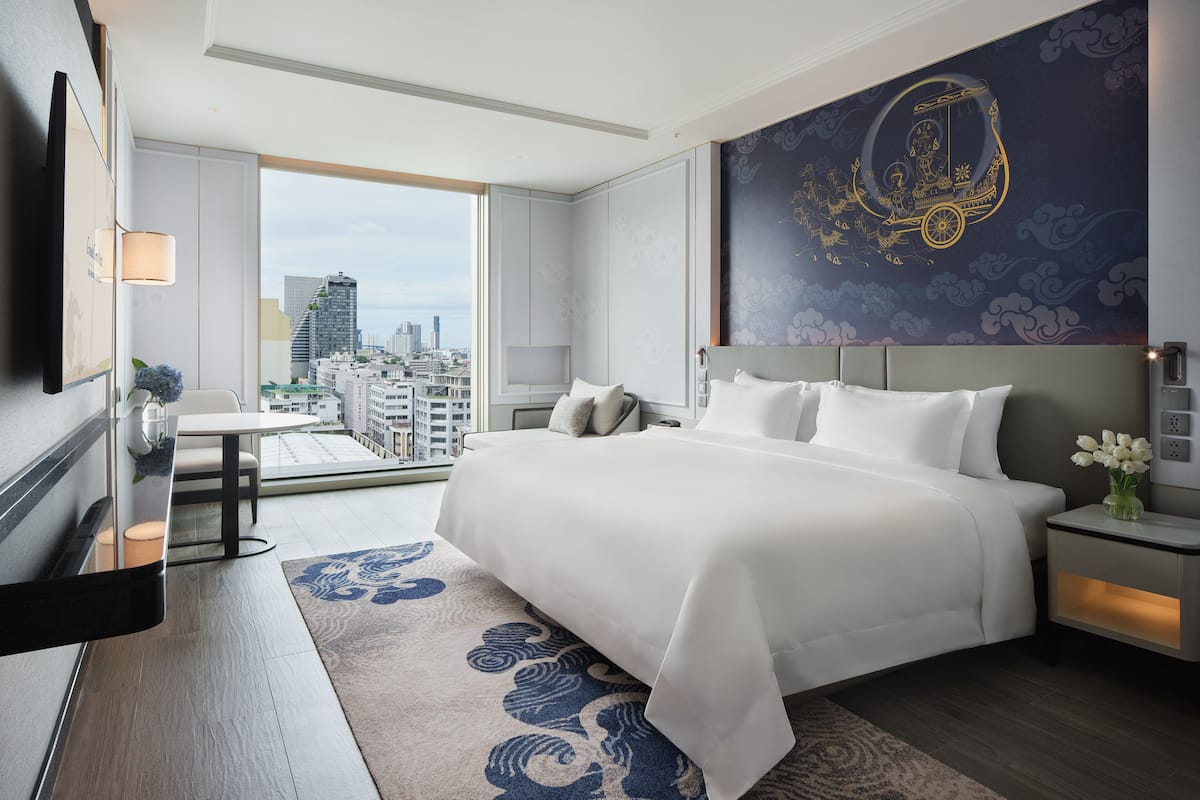 A modern hotel room with a large bed, a city view, a decorative wall mural, and a small table with a vase.