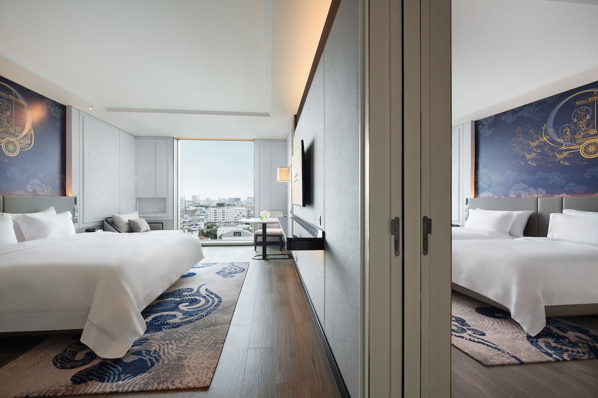 A modern hotel room with two beds, a desk, and a view of the city.