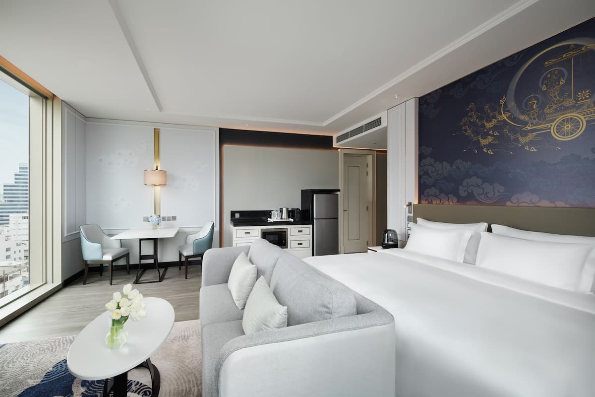 A modern hotel room with a large bed, a sofa, a dining table, and a kitchen area.