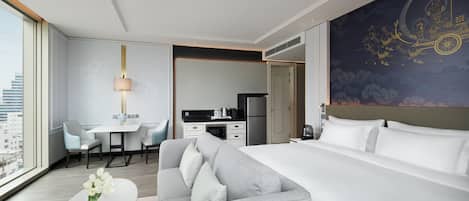 A modern hotel room with a large bed, a sofa, a dining table, and a kitchen area.