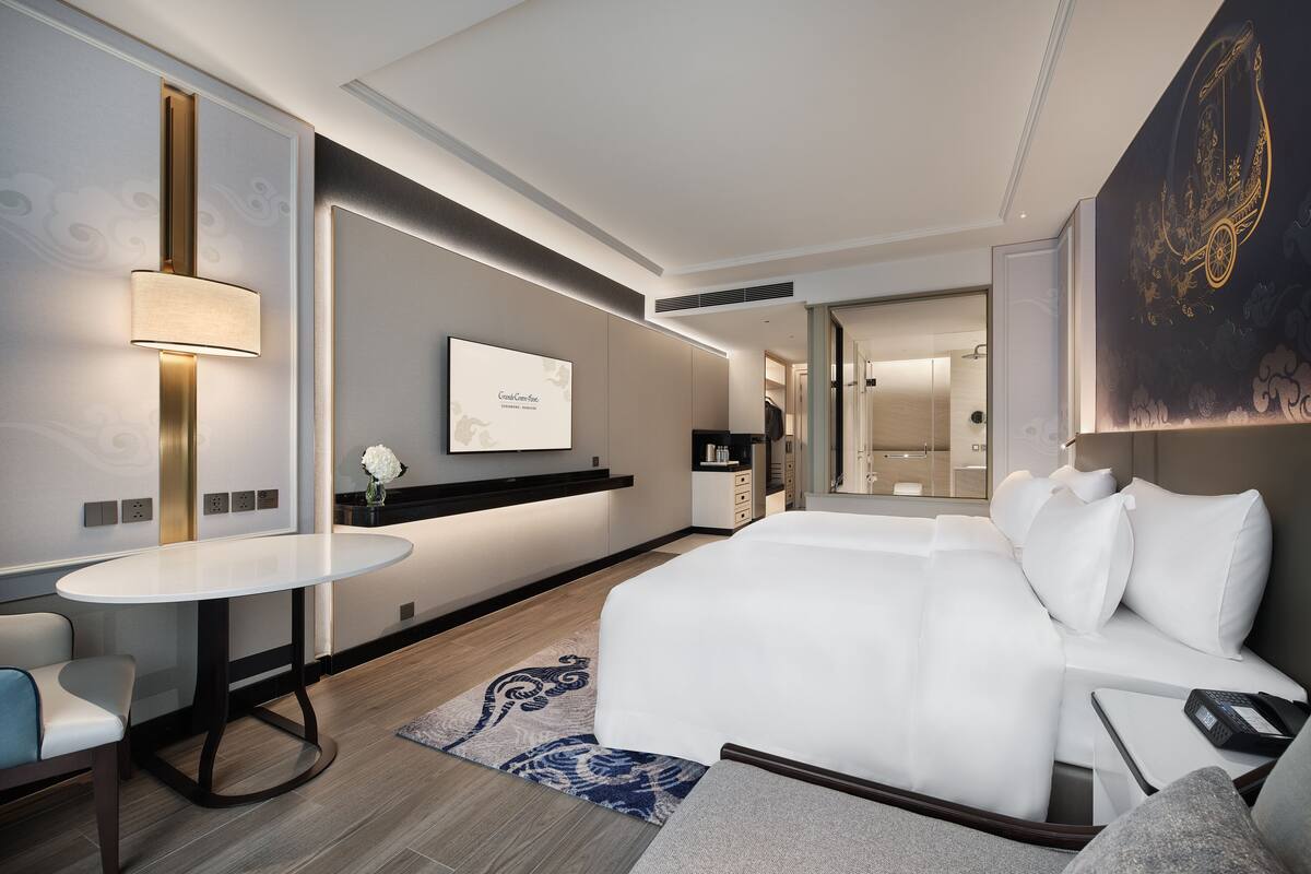 A modern hotel room with a large bed, a bedside table, a TV, and a seating area.
