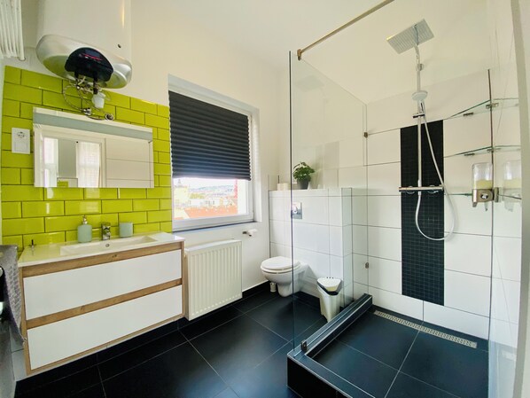 Une salle de bain moderne avec un mur recouvert de carreaux jaunes, un lavabo blanc avec un miroir, des toilettes et une douche séparée par une paroi en verre.