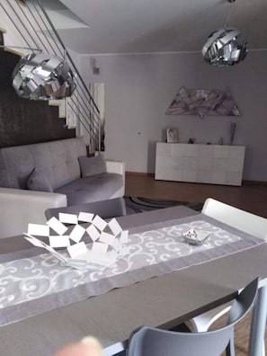 A modern living room with a grey sofa, a dining table with a patterned table runner, and a staircase with a metal railing. - 2 Bedroom Villa in Castellammare del Golfo (Castellammare del Golfo)