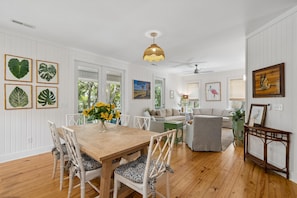 Un espace repas avec une table en bois et des chaises blanches, un ventilateur de plafond et des œuvres d’art accrochées au mur.
