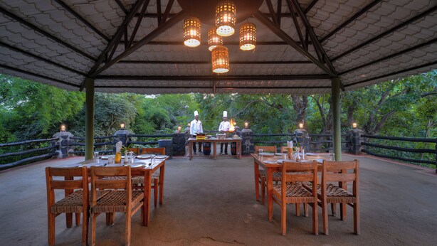Un espacio de comedor al aire libre con mesas y sillas de madera, techo cubierto y luces colgantes.