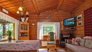 Et hyggeligt interiør i en hytte med trælofter, et fjernsyn monteret på væggen, en sofa, en lænestol, et bord med blomstret dug og et skab med glaslåger.