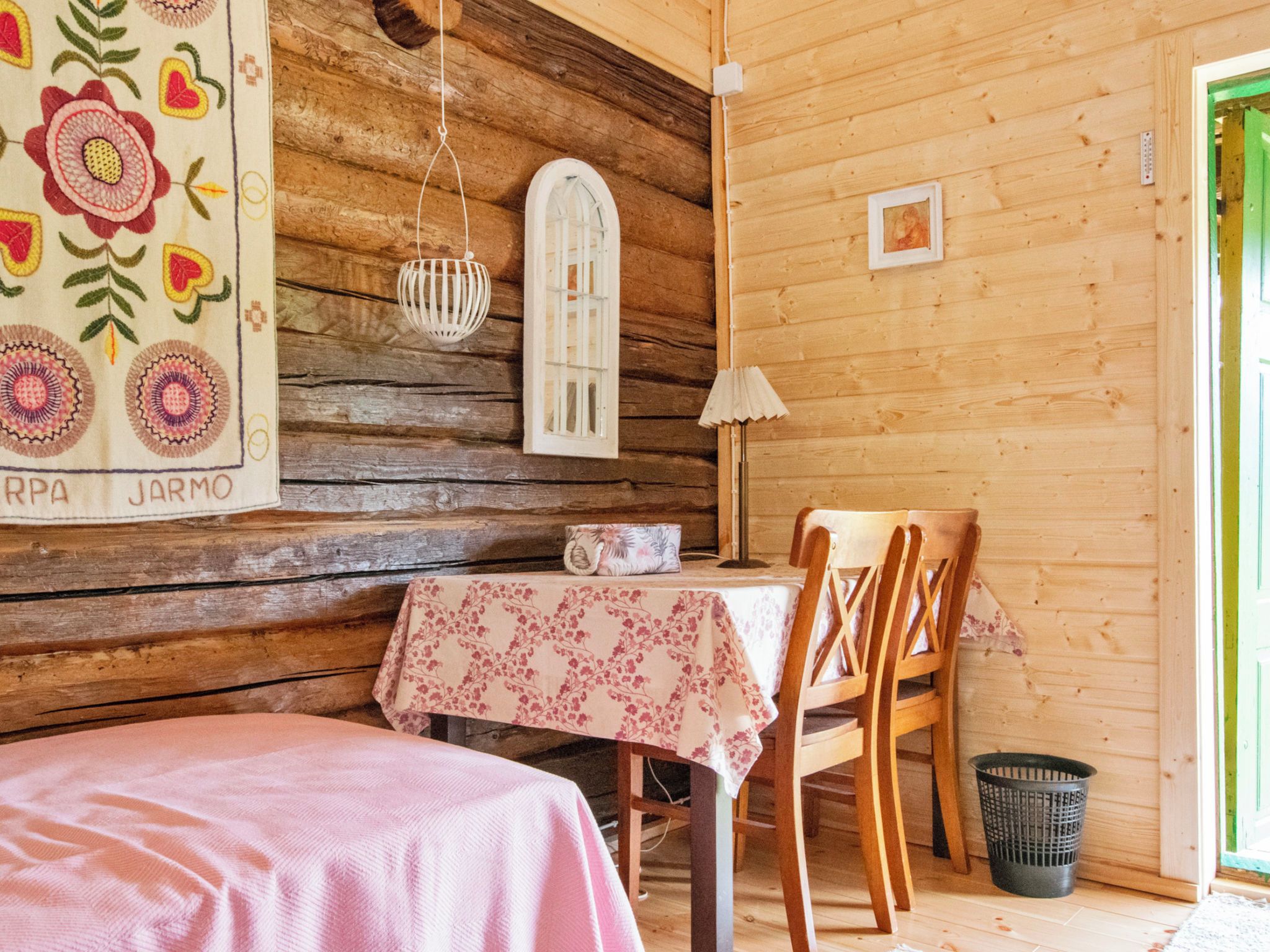 A cozy cabin interior with a wooden wall, a table with a floral tablecloth, a chair, a hanging light fixture, and a small window.