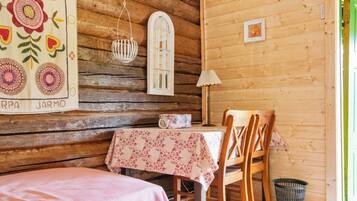 A cozy cabin interior with a wooden wall, a table with a floral tablecloth, a chair, a hanging light fixture, and a small window.