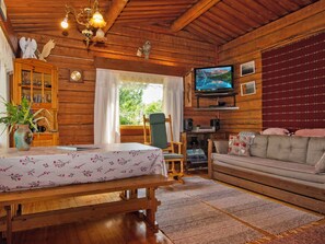 Interior acogedor de una cabaña con techo de madera, un televisor en la pared, un sofá, una mesa con mantel floreado y una ventana con cortinas.