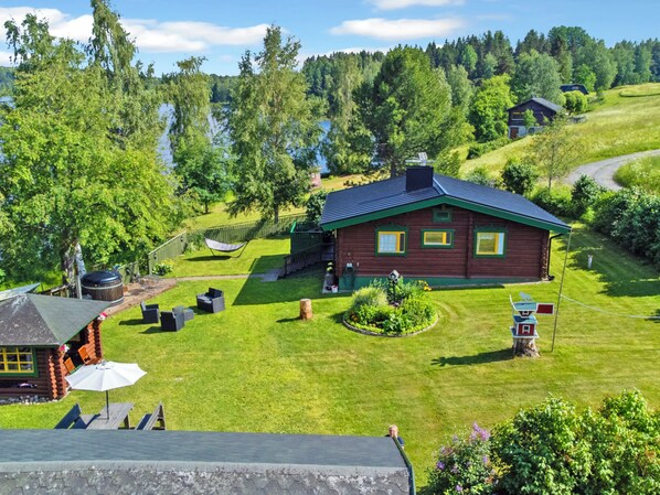 A cabin with a grassy yard, a pond, and a forested hill in the background.