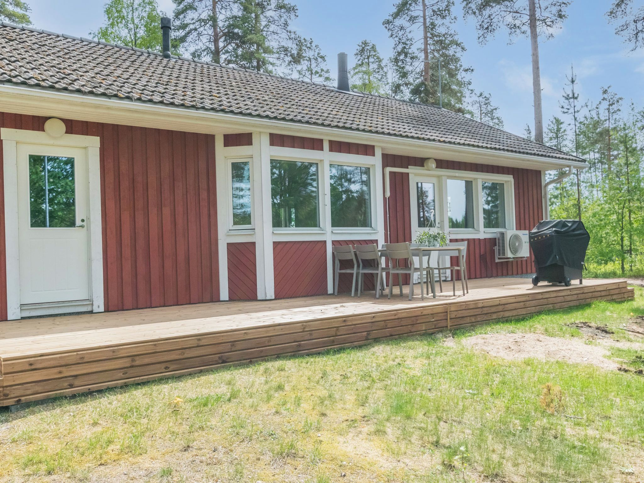 A single-story building with a red exterior, white trim, and a tiled roof. There is a covered outdoor seating area with a table and chairs, and a grill.