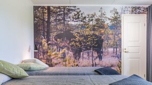 Ein Schlafzimmer mit einer großen Wandmalerei zum Thema Wald, einem Bett mit grauer Bettwäsche und einer Tür rechts.